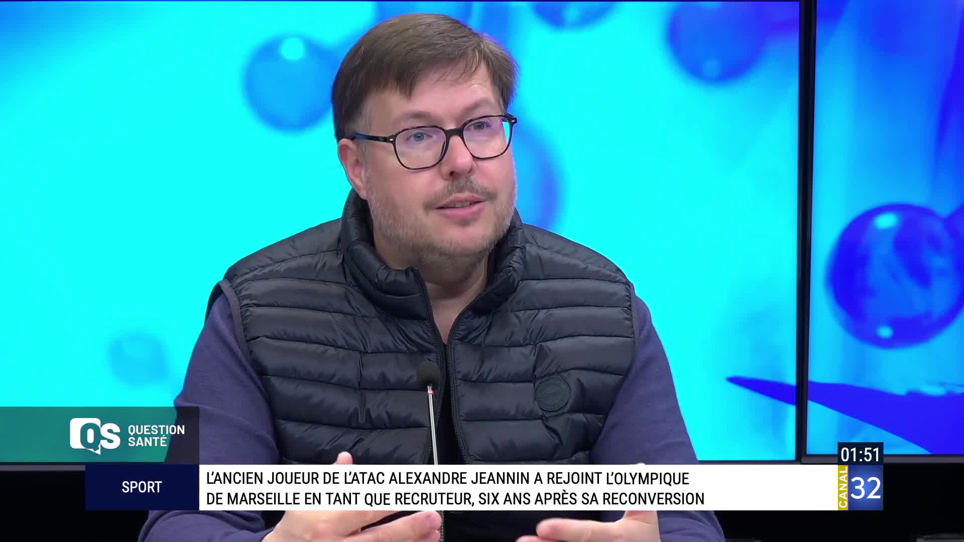 A man with glasses speaks into a microphone, his vest zipped over a purple shirt. Behind him, a bright blue screen displays abstract shapes, and a graphic at the bottom of the screen reads "Canal 32." A man with glasses speaks into a microphone, his vest zipped over a purple shirt. Behind him, a bright blue screen displays abstract shapes, and a graphic at the bottom of the screen reads "Canal 32."