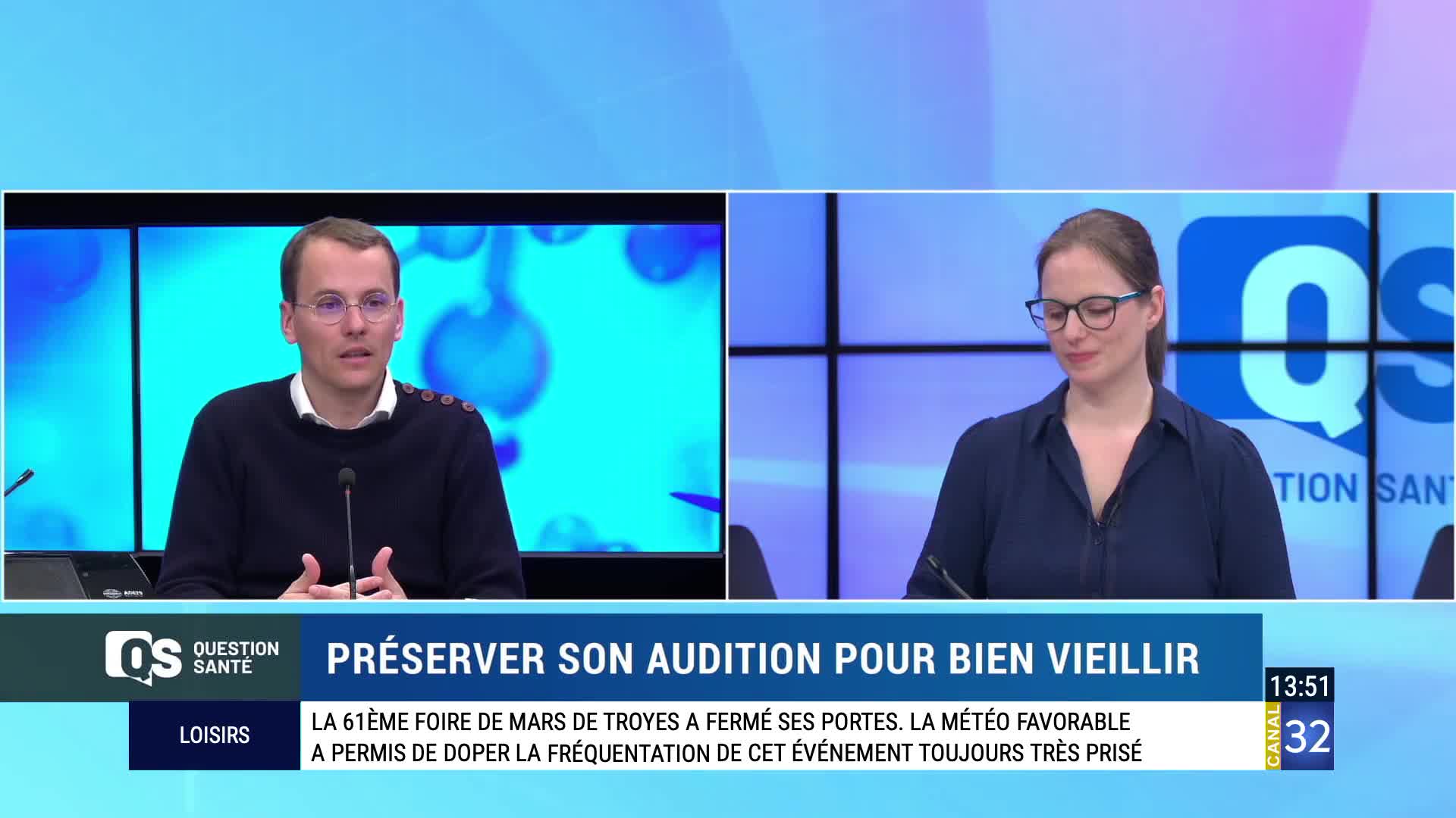 A man in a dark sweater speaks into a microphone, his hands gesturing slightly. Beside him, a woman in a navy blouse looks down, her glasses catching the light. The Canal 32 logo appears on a screen behind them. A man in a dark sweater speaks into a microphone, his hands gesturing slightly. Beside him, a woman in a navy blouse looks down, her glasses catching the light. The Canal 32 logo appears on a screen behind them.