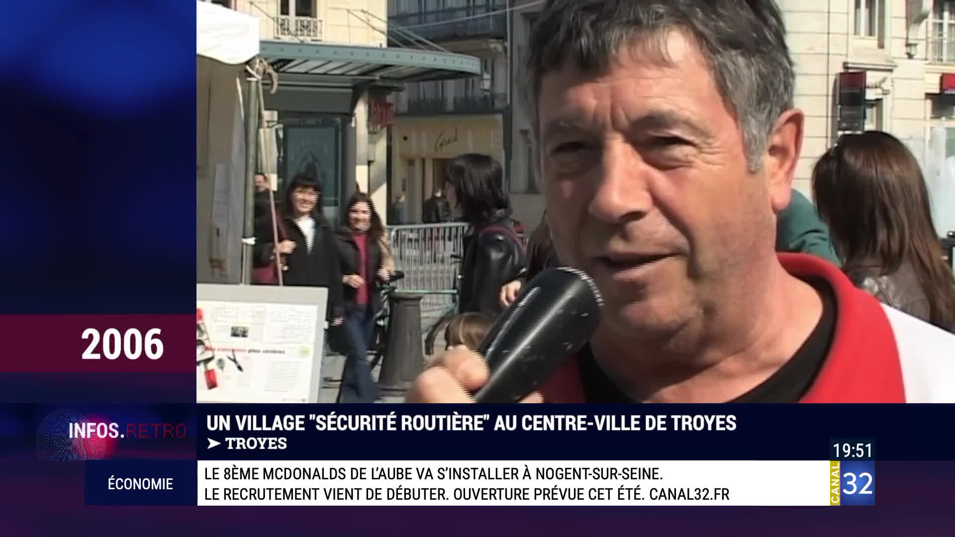 A man holds a microphone, speaking into it as two women walk past in the background. The scene unfolds on a sunny day in Troyes, France. A man holds a microphone, speaking into it as two women walk past in the background. The scene unfolds on a sunny day in Troyes, France.