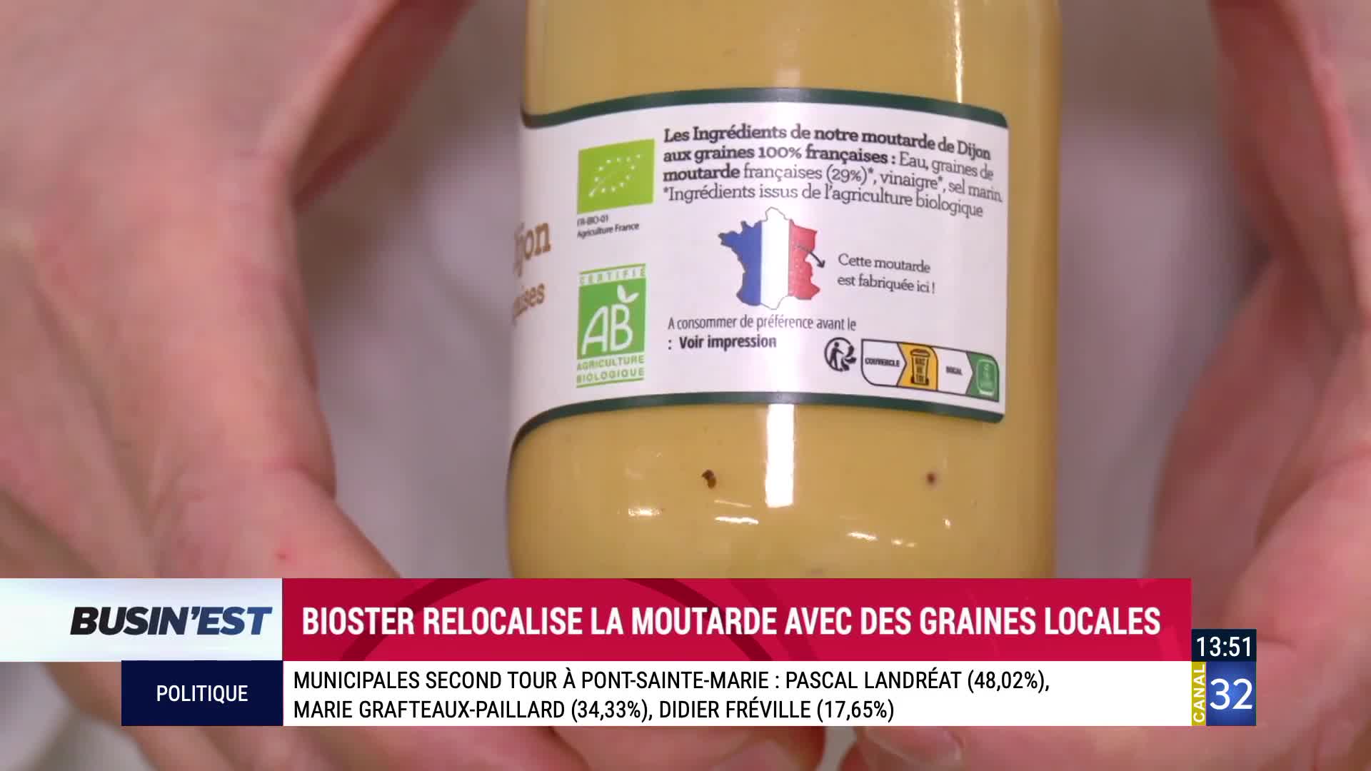 A hand holds a jar of Dijon mustard, its label detailing French ingredients and organic certification. A lower third on Canal 32 reports on local mustard production and municipal election results. A hand holds a jar of Dijon mustard, its label detailing French ingredients and organic certification. A lower third on Canal 32 reports on local mustard production and municipal election results.
