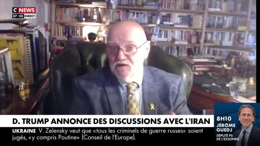 A man in a suit, with a yellow ribbon pinned to his lapel, is speaking on CNews. Behind him, a wall of books fills the space, and a lower screen shows another man's face.
