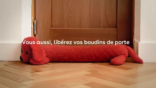 A long, red, stuffed dog lies on the wooden floor, its head resting against a white baseboard. The dog's body stretches across the bottom of a closed wooden door, its tail disappearing out of frame.