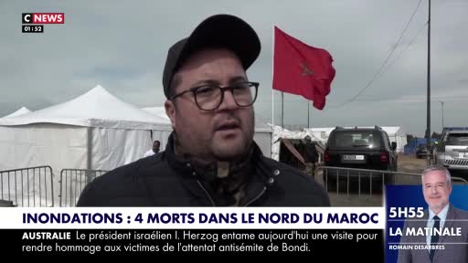A man in a black baseball cap and glasses speaks directly to the camera. Behind him, a large red Moroccan flag flutters against a cloudy sky, with white tents and vehicles suggesting an outdoor event or relief effort.