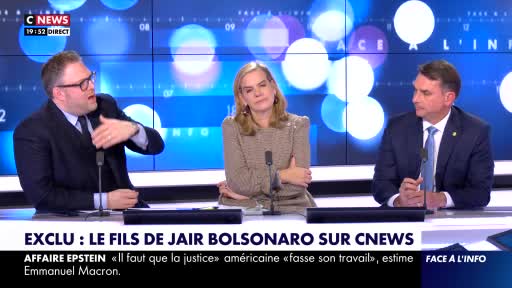 A man in a suit gestures emphatically while speaking into a microphone. To his right, a woman in a tweed jacket listens intently, arms crossed. Further right, another man in a suit looks thoughtfully to the side.