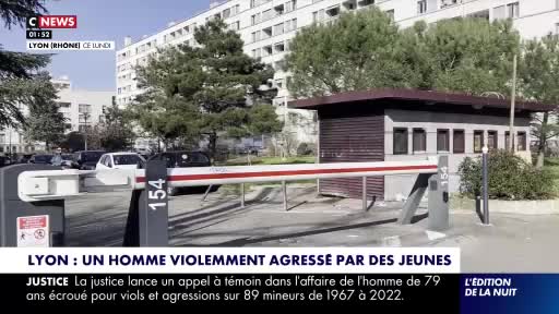 A white car is slowly passing through a raised barrier at the entrance to a parking lot. Behind it, a small security booth stands next to a tall apartment building.