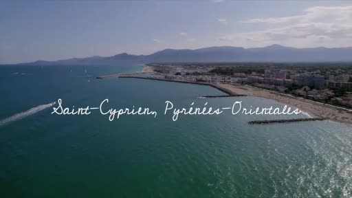 A small boat speeds across the turquoise water, leaving a white wake behind it. Beyond, the coast of Saint-Cyprien in France stretches out, with a town and sandy beaches meeting the Mediterranean.