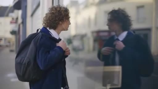 A young man with curly hair, wearing a school uniform and backpack, pauses by a shop window in a French town. His reflection gazes back at him as he adjusts his jacket.