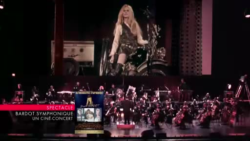 An orchestra is playing on stage, illuminated by spotlights. Above them, a large screen displays Brigitte Bardot on a motorcycle. An orchestra is playing on stage, illuminated by spotlights. Above them, a large screen displays Brigitte Bardot on a motorcycle.