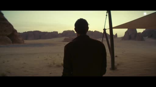 A man stands silhouetted against the pale desert sky, facing a rugged landscape. A makeshift shelter with a tan tarp stretches out beside him. A man stands silhouetted against the pale desert sky, facing a rugged landscape. A makeshift shelter with a tan tarp stretches out beside him.