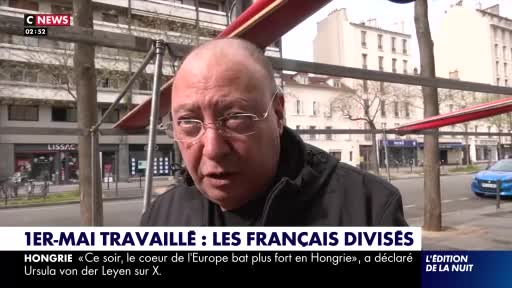 A man speaks directly to the camera, his glasses reflecting the Parisian street behind him. A blue car drives past on the road. A man speaks directly to the camera, his glasses reflecting the Parisian street behind him. A blue car drives past on the road.