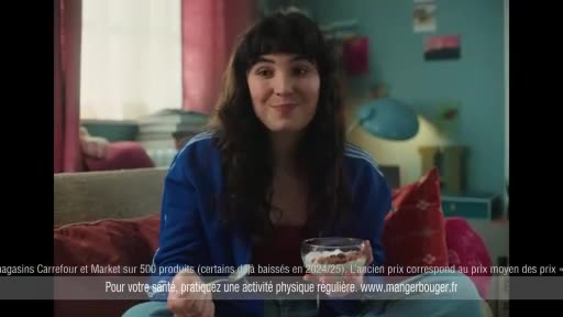 A young woman with dark hair and bangs smiles, holding a bowl of dessert. She's wearing a blue track jacket over a red top, relaxing on a couch.