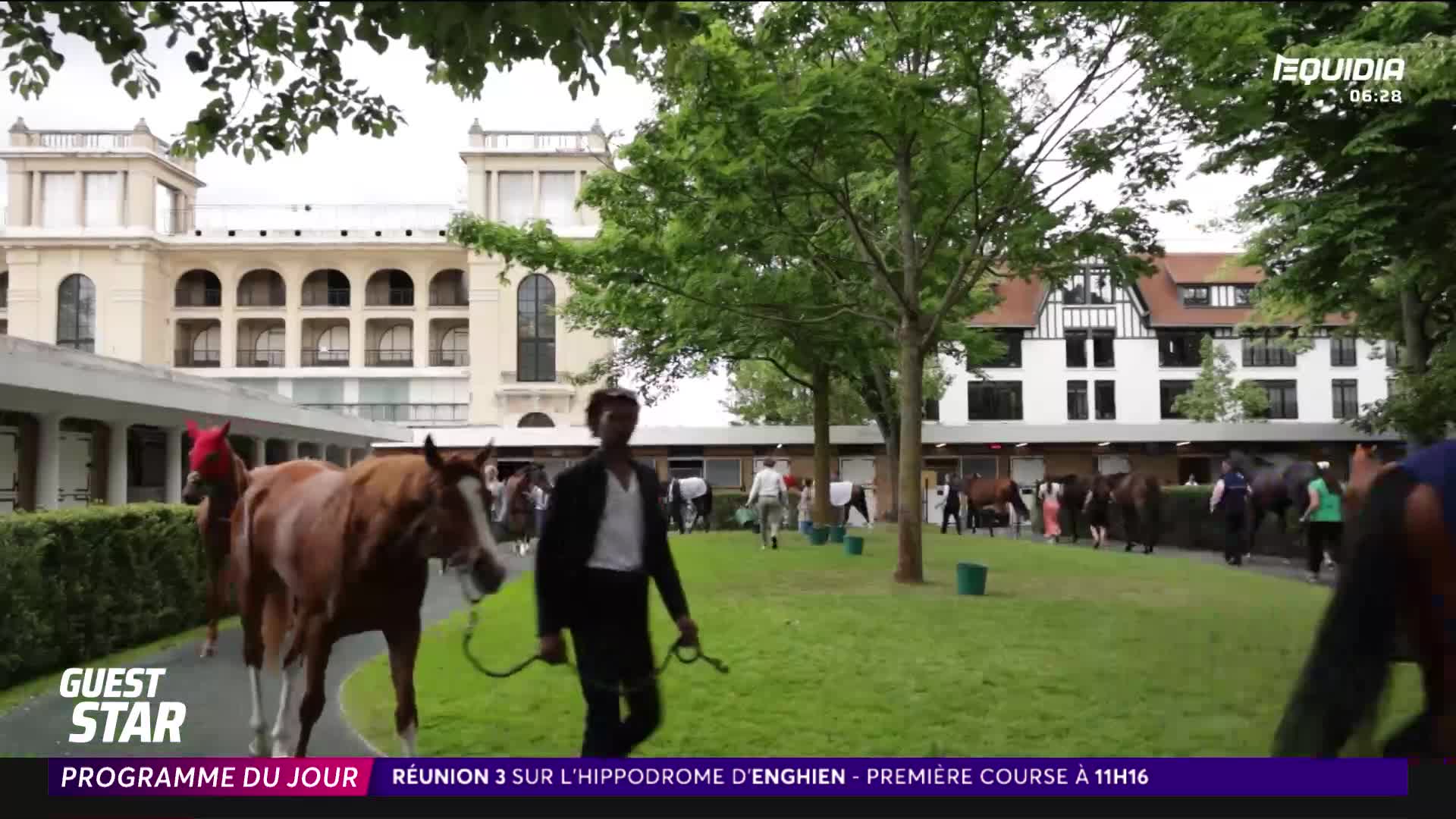 A handler leads a chestnut horse, its head covered in a red hood, past the stables. Other horses and people are milling about, getting ready for the day's races at the Hippodrome d'Enghien, as shown on Equidia.
