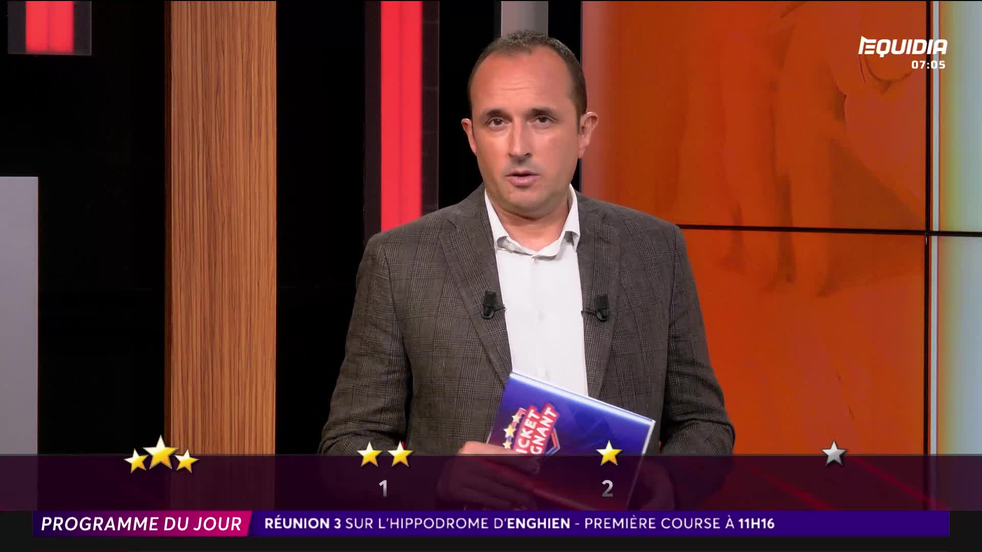 A man in a grey jacket is holding a book, speaking directly to the camera on Equidia. The screen shows a program guide for a race at the Hippodrome d'Enghien in France.
