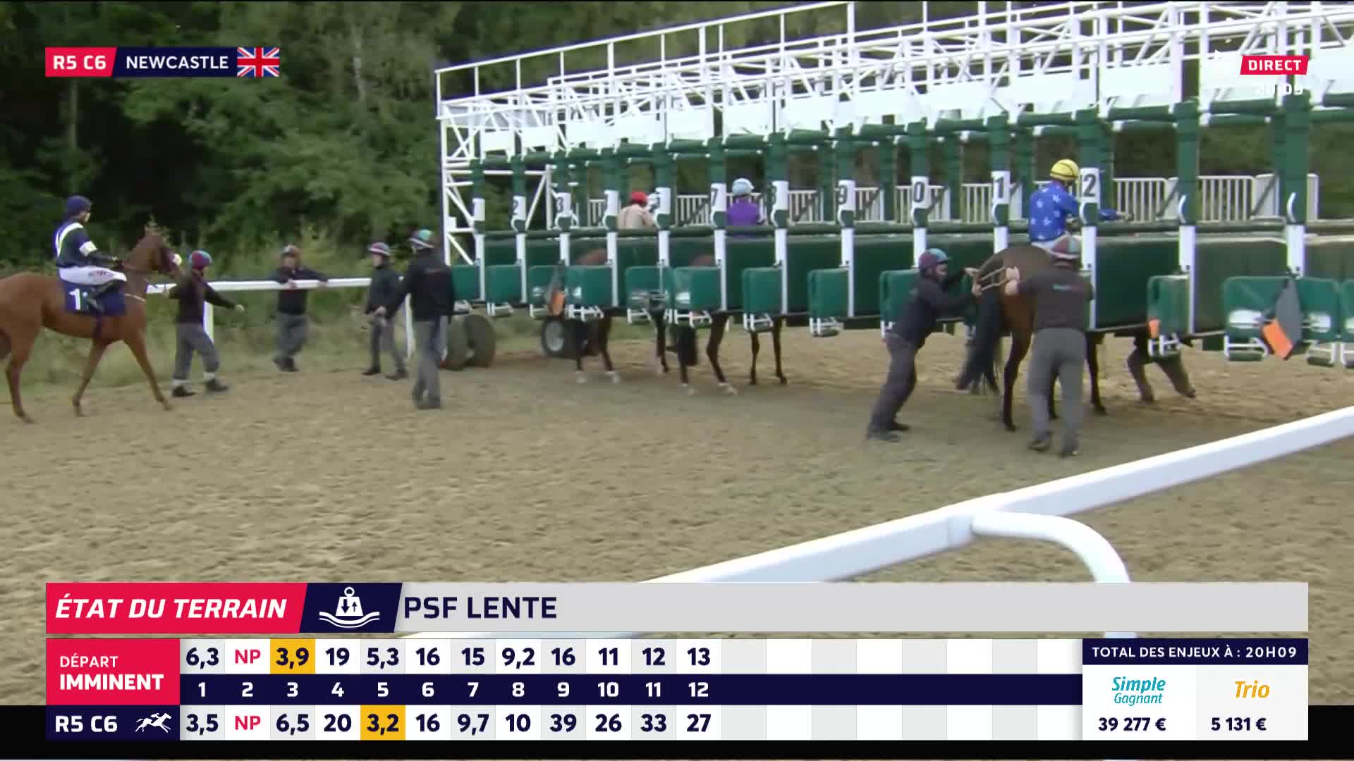 Horses are being guided into the starting gates, with handlers steadying them. The jockeys are already mounted and ready to go.
