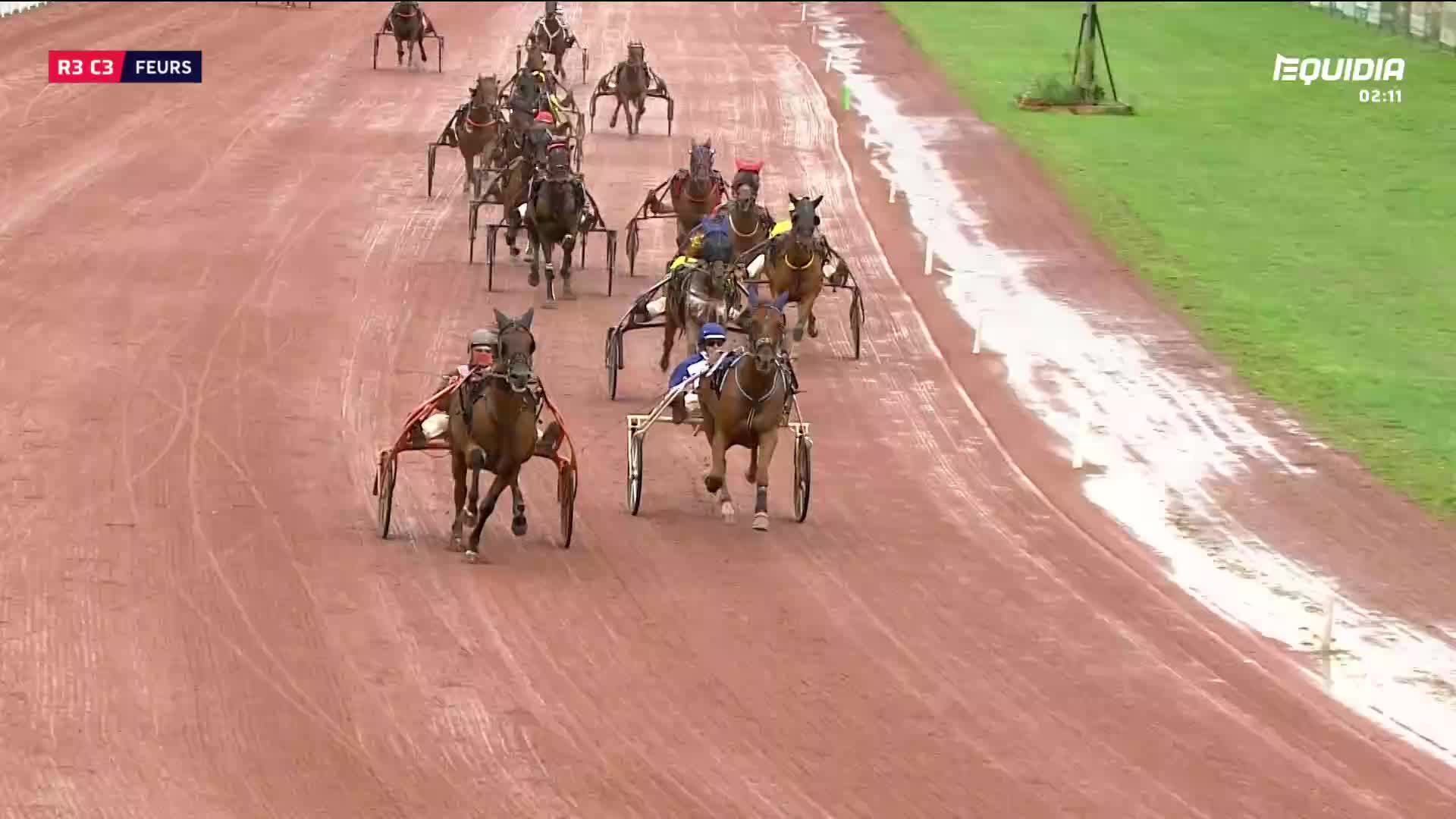 The horses, pulling sulkies, thunder down the track, their hooves kicking up the reddish dirt. The Equidia broadcast is showing a race in France, with the lead horse straining ahead.
