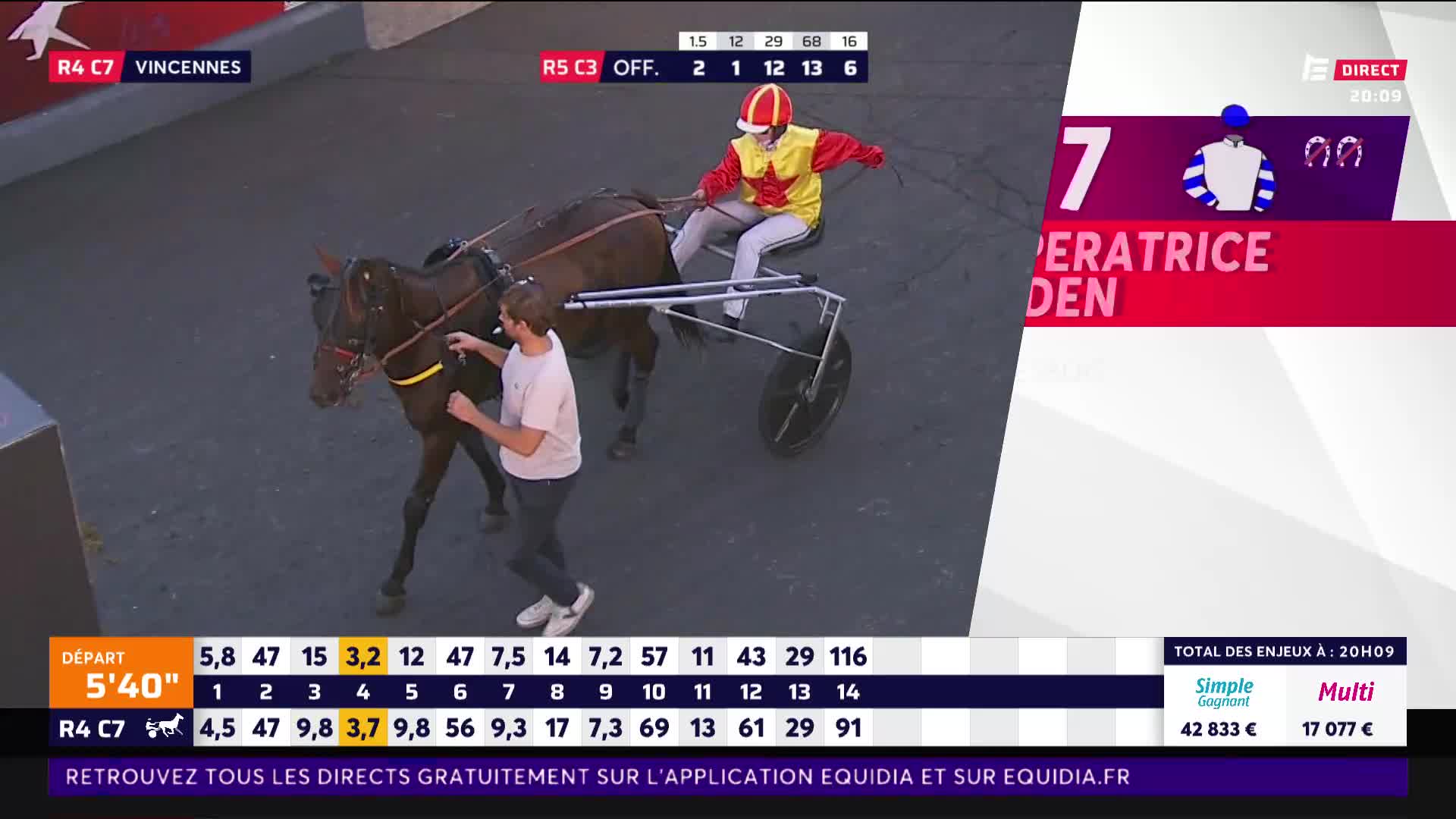 A man in a red and yellow racing suit sits in a sulky behind a trotting horse. Another man in a light pink shirt walks alongside the horse, guiding it.
