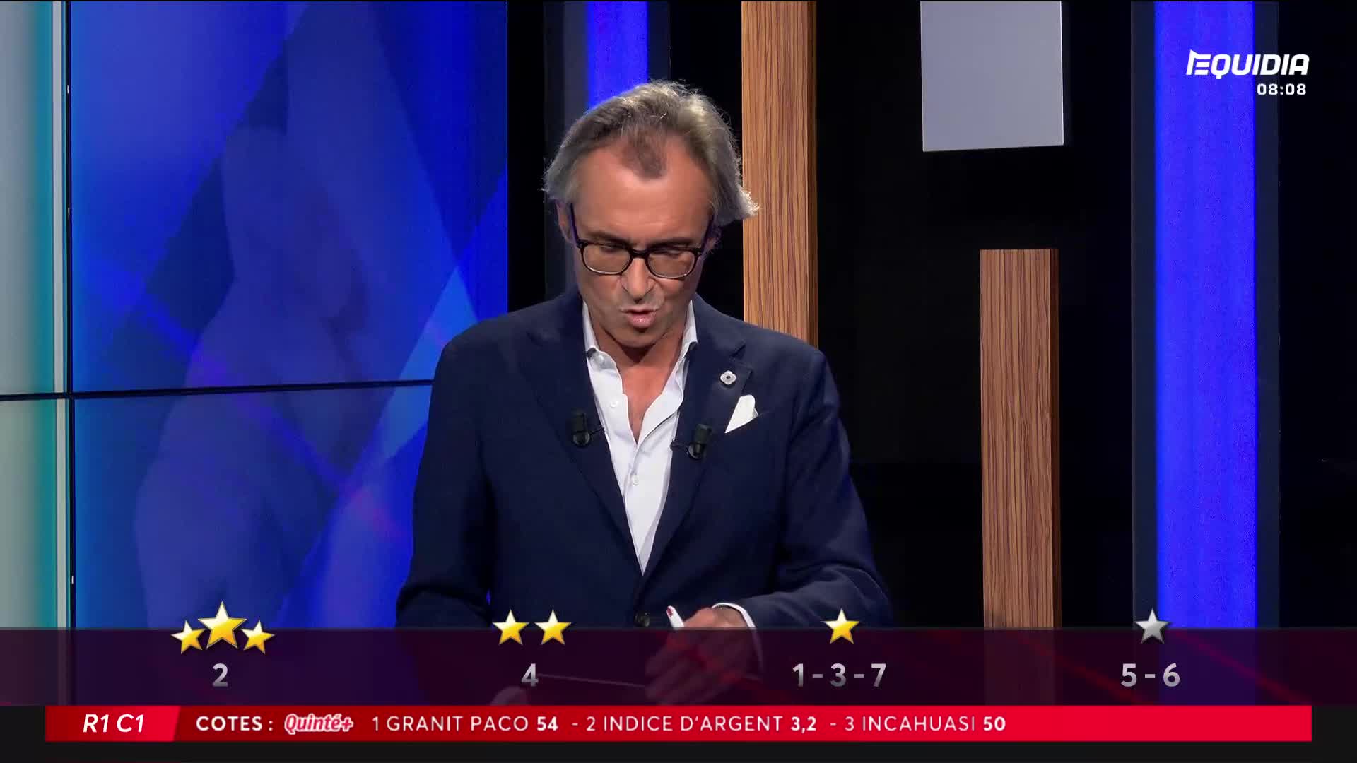 A man in a navy blazer and glasses speaks intently, his gaze fixed on something just out of view. Below him, the Equidia screen displays odds and race information.
