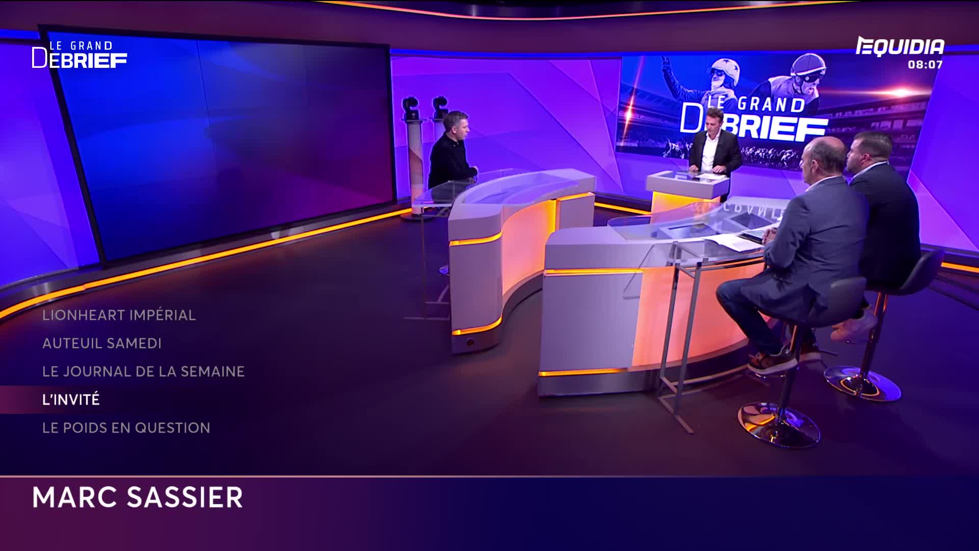 Three men sit at a curved desk, discussing something on Equidia's "Le Grand Débrief" set. A large screen behind them displays the show's logo and graphics.

