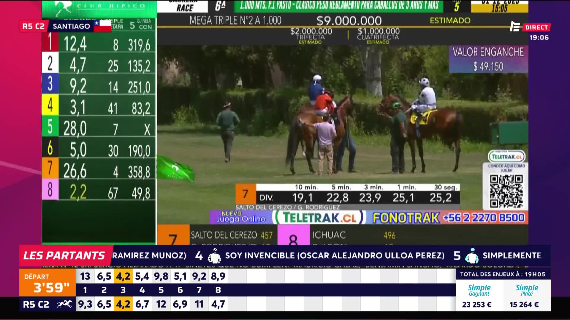 Jockeys on horseback are being led onto the track, with handlers walking alongside. The horses, in their racing colors, are grouped near the starting gate as the race information is displayed on the screen.
