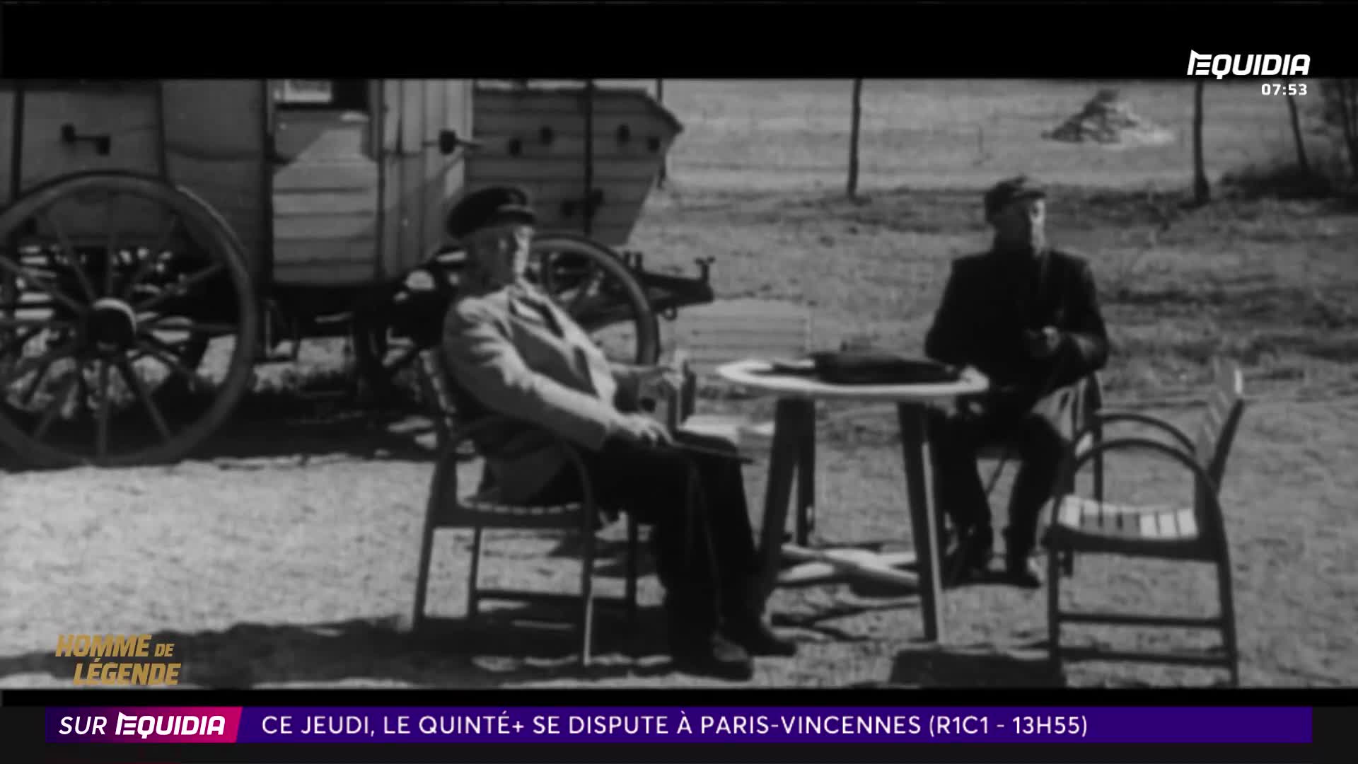 Two men sit outdoors near a horse-drawn wagon. One man, wearing a cap and jacket, relaxes in a chair while the other, in a dark suit, sits at a table.