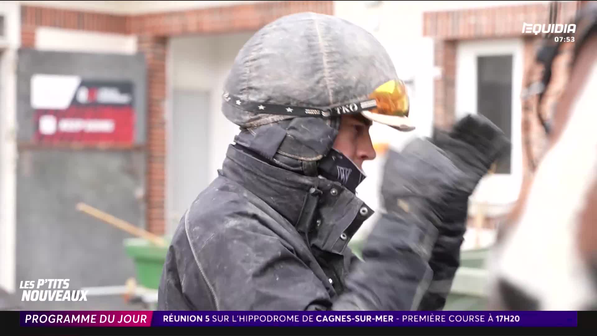 A jockey adjusts his helmet, the goggles reflecting the overcast sky. Another horse, its flank a blur of white, stands close by.
