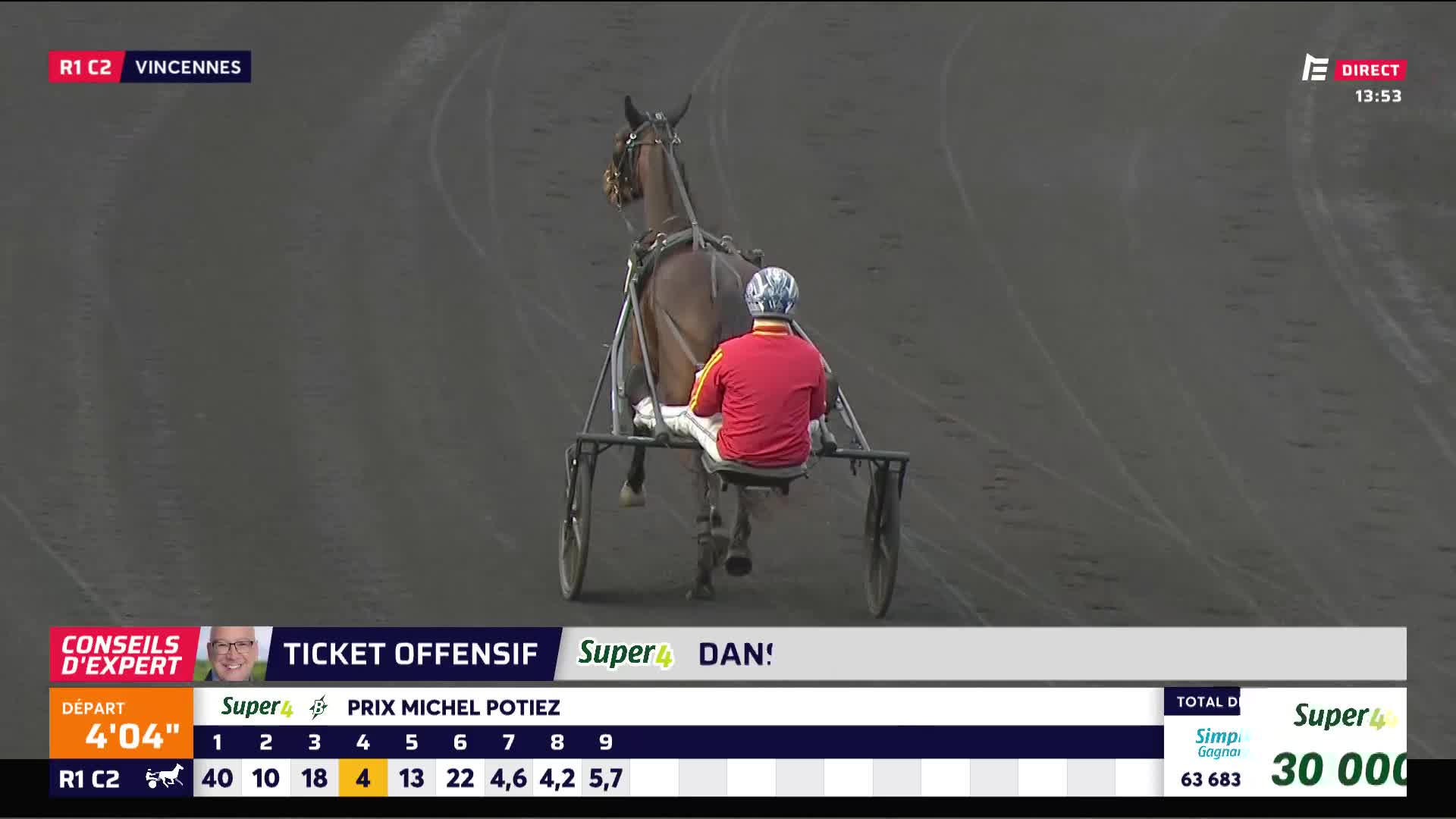 A trotter pulls its sulky with a driver in a red shirt and blue helmet across the Vincennes track. The horse's hooves kick up dust as it moves steadily forward.