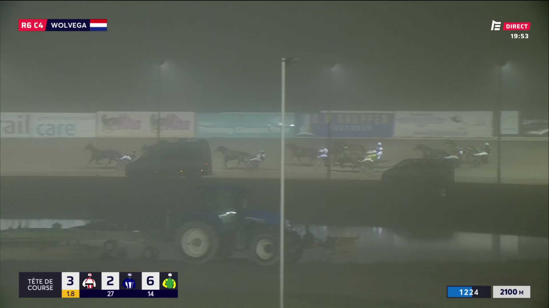 The horses are trotting down the track in the dim light, their sulkies kicking up dust. A tractor sits idle in the foreground, its blue paint barely visible through the haze.
