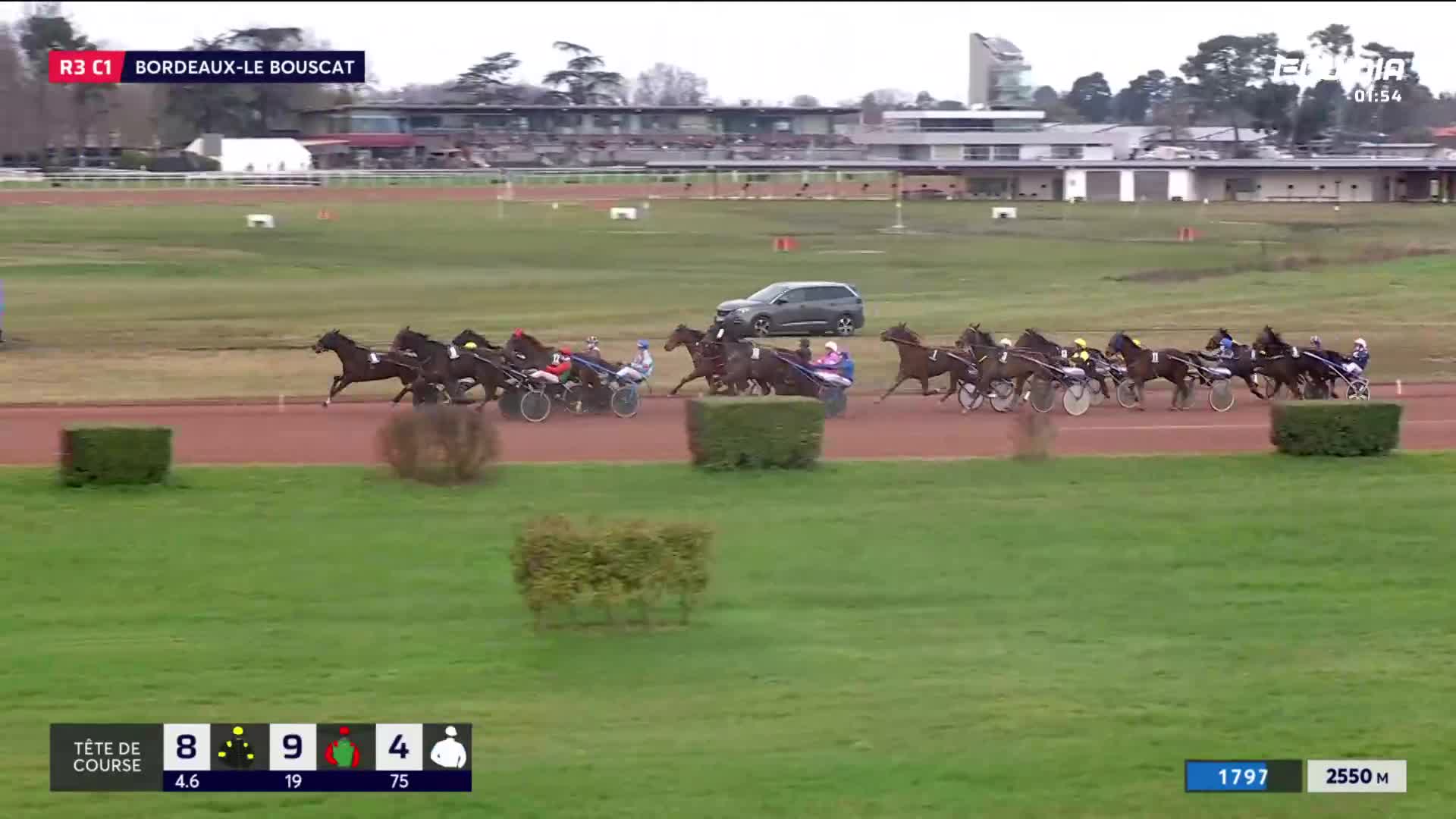 The horses are thundering down the track at Bordeaux-Le Bouscat, their sulkies kicking up dust. A grey car follows closely behind the leading pack.