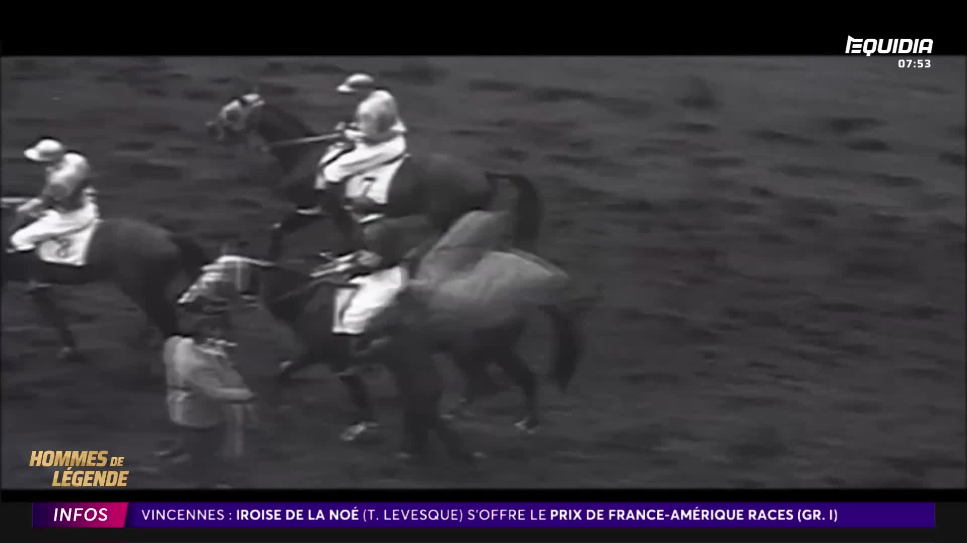 The horses surge forward, their riders leaning low.  One jockey in a white cap and silks is clearly ahead, pushing his mount.