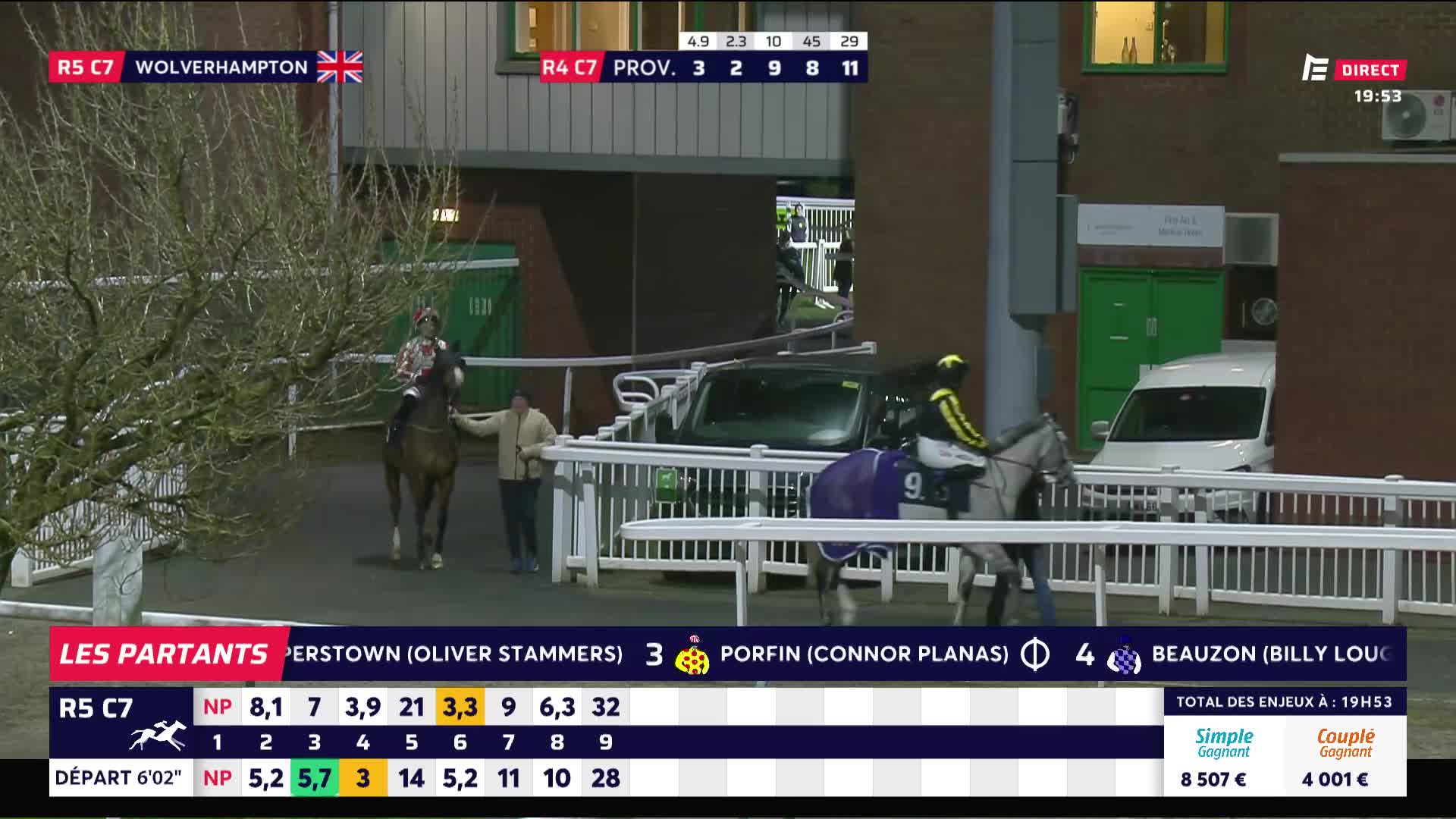 A handler leads a chestnut horse and rider past a white railing. Another horse, this one grey with a purple blanket, is being led by a jockey in yellow and black silks.