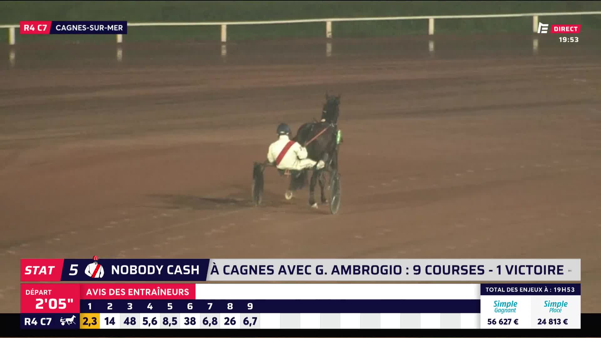A dark horse pulls a sulky with a driver in a white and red uniform down the dirt track at Cagnes-sur-Mer. The horse's hooves kick up a spray of fine sand as it moves steadily forward.