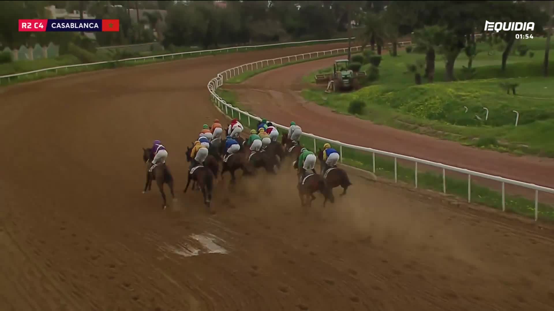 The horses are thundering down the track, kicking up a cloud of dust as they round the bend. A jockey in purple and white pulls slightly ahead of the pack. The horses are thundering down the track, kicking up a cloud of dust as they round the bend. A jockey in purple and white pulls slightly ahead of the pack.