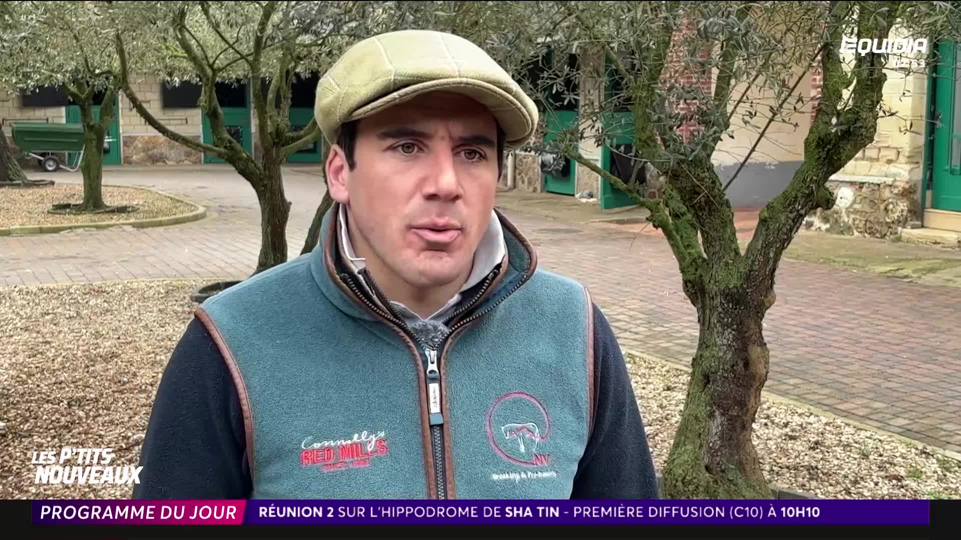 A man in a flat cap and a teal vest speaks directly to the camera. Behind him, a trailer is parked near a stable building.