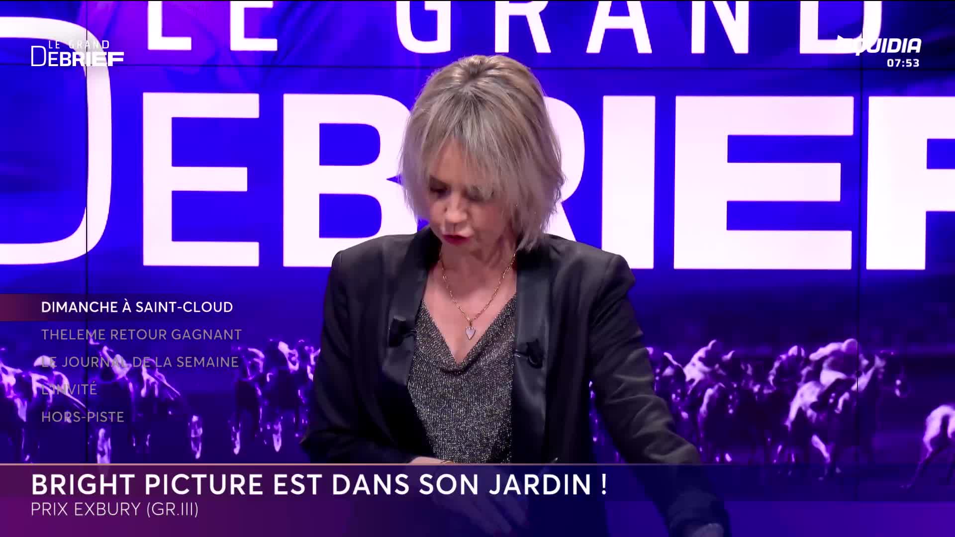 A woman in a dark jacket stands before a large screen displaying "LE GRAND DEBRIEF" in bold white letters. Below her, a line of horses gallops across the screen, their forms silhouetted against a vibrant purple background.