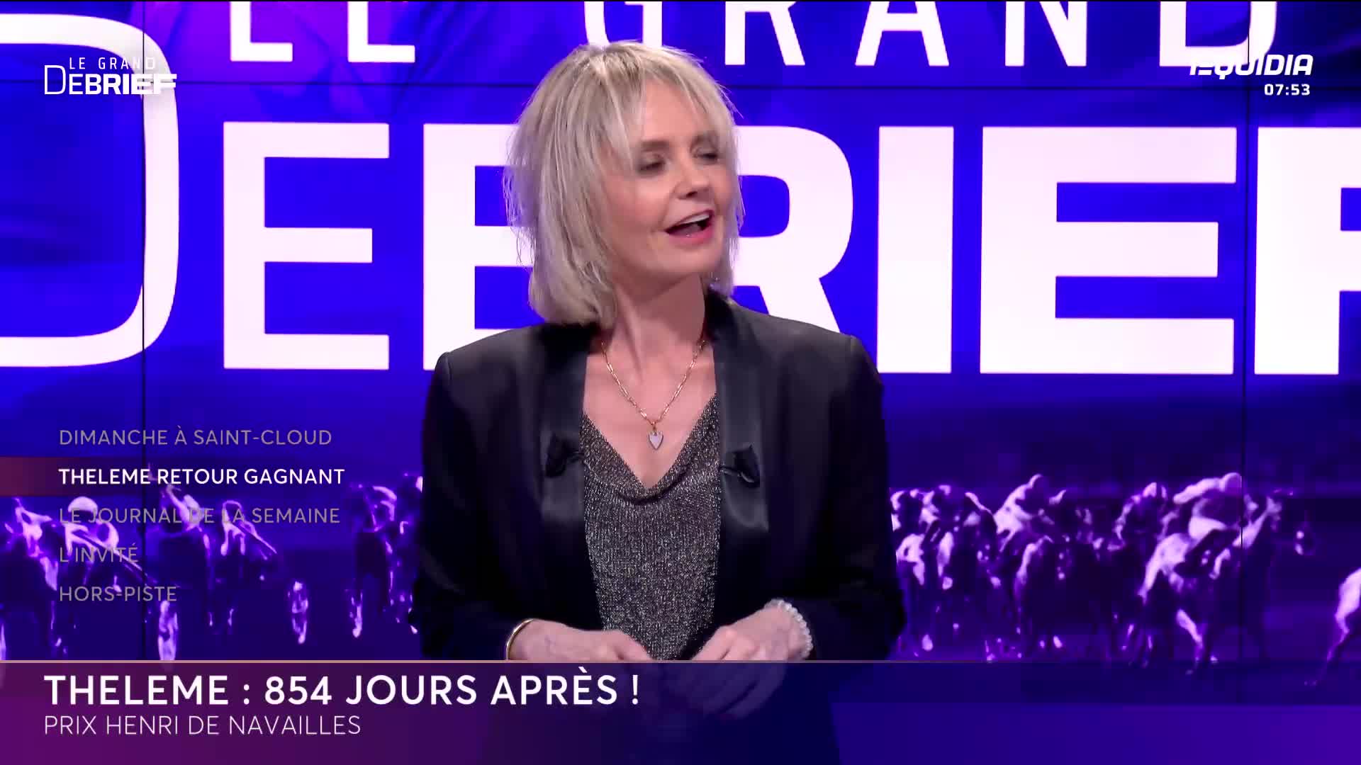 A woman in a black jacket speaks animatedly, her blonde hair framing her face. Behind her, a graphic displays "LE GRAND DEBRIEF" in large white letters, with a row of horses running across the bottom.