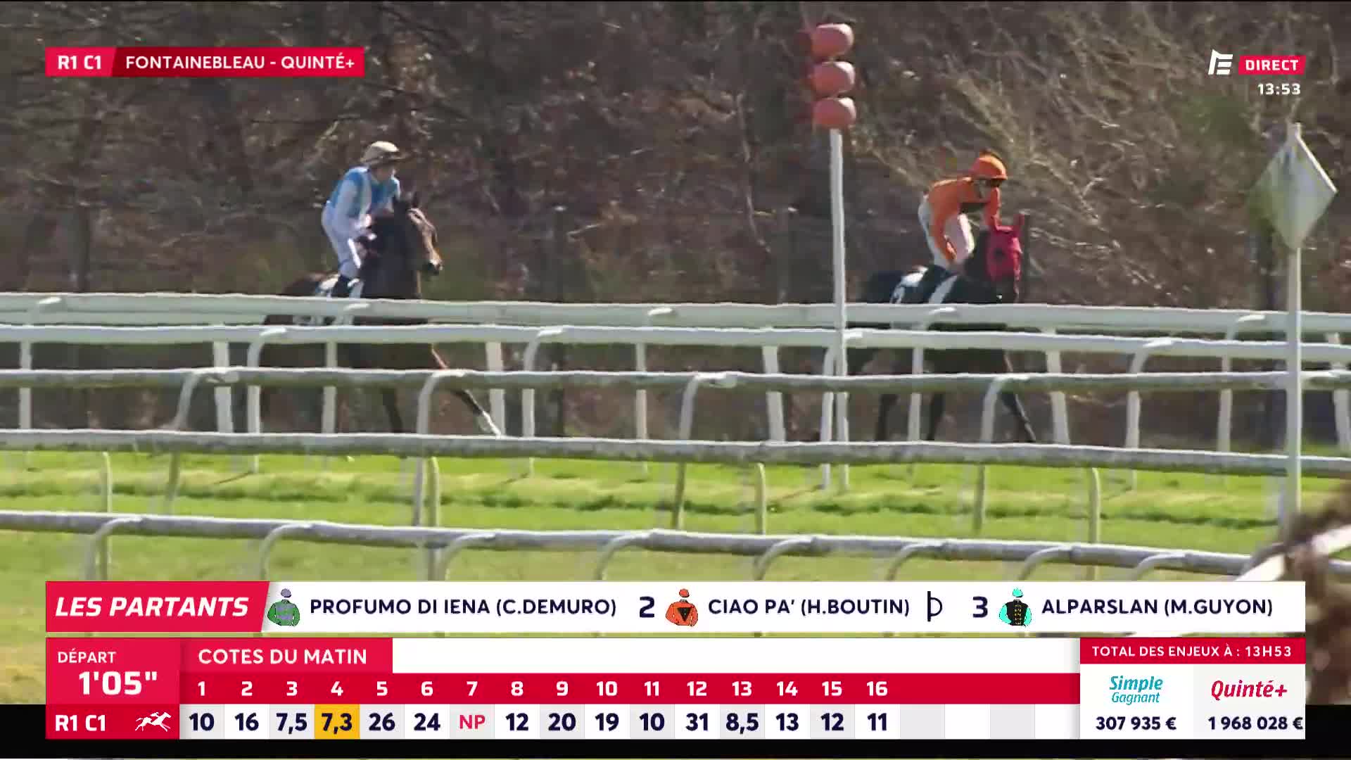A jockey in a pale blue jacket guides his horse past the white rails. Another rider, in an orange top, is a few lengths behind, leaning forward over his mount.