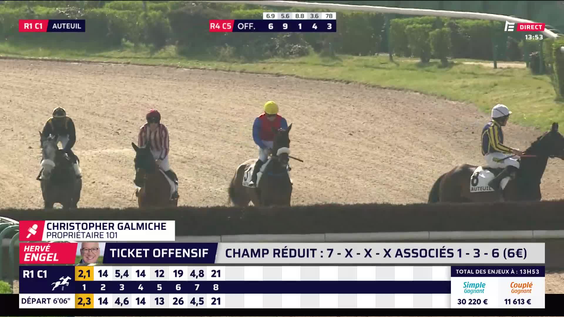 Four horses and their jockeys are approaching a fence, their strides steady on the dirt track. The lead horse, ridden by a jockey in a yellow helmet, is just about to clear the obstacle.