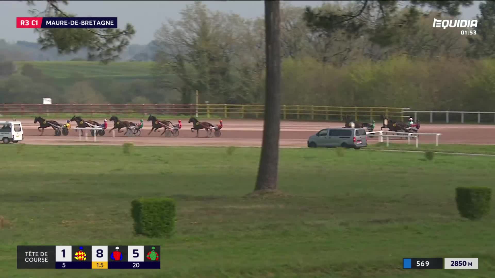 The horses are pulling their sulkies down the track at Maure-de-Bretagne. A few cars follow behind, keeping pace with the race.