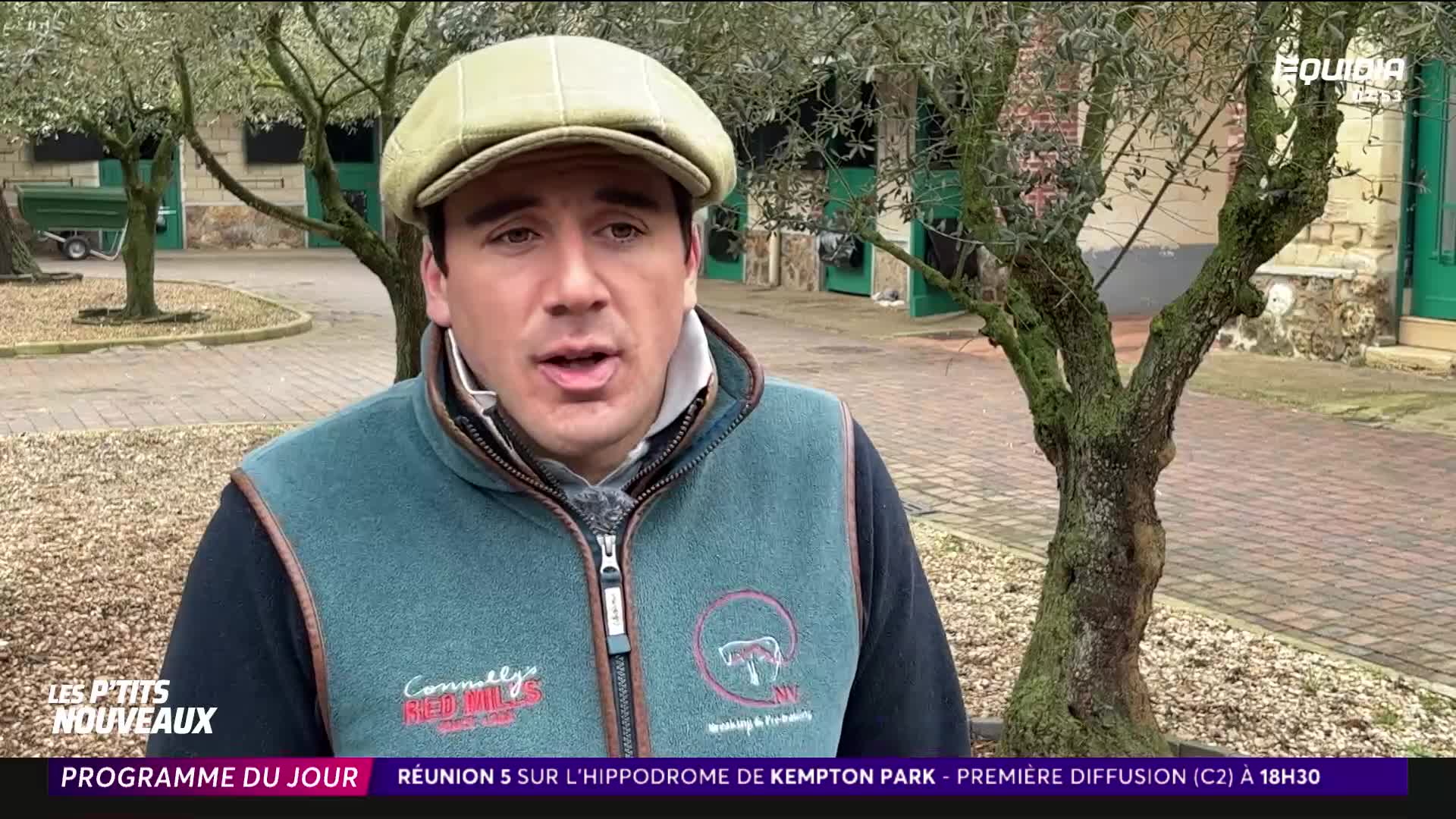A man in a flat cap and gilet speaks directly to the camera. Behind him, stable doors stand open, and a small green trailer is parked near a young tree.