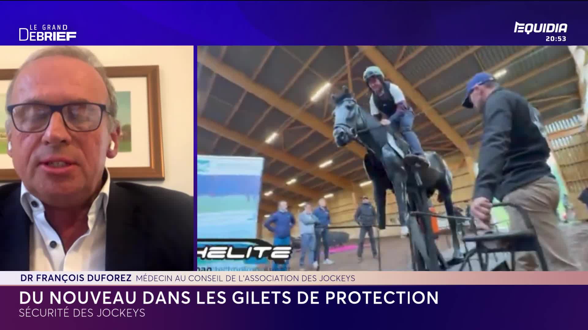 A jockey sits atop a dark horse, their helmet and vest clearly visible. A man in a blue cap steadies the horse's bridle, while others observe in the background of this indoor training facility. A jockey sits atop a dark horse, their helmet and vest clearly visible. A man in a blue cap steadies the horse's bridle, while others observe in the background of this indoor training facility.