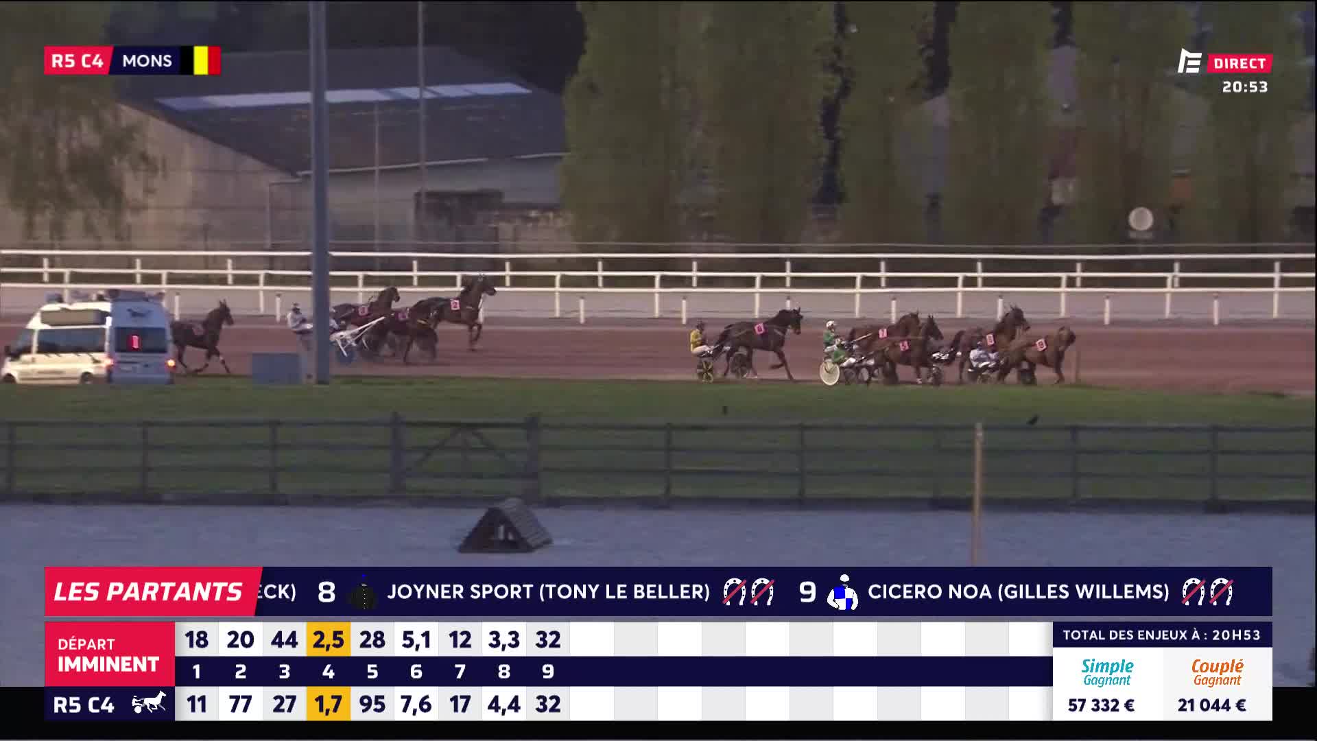 The horses are pulling their sulkies around the track at Mons. A white van is parked near the starting line. The horses are pulling their sulkies around the track at Mons. A white van is parked near the starting line.
