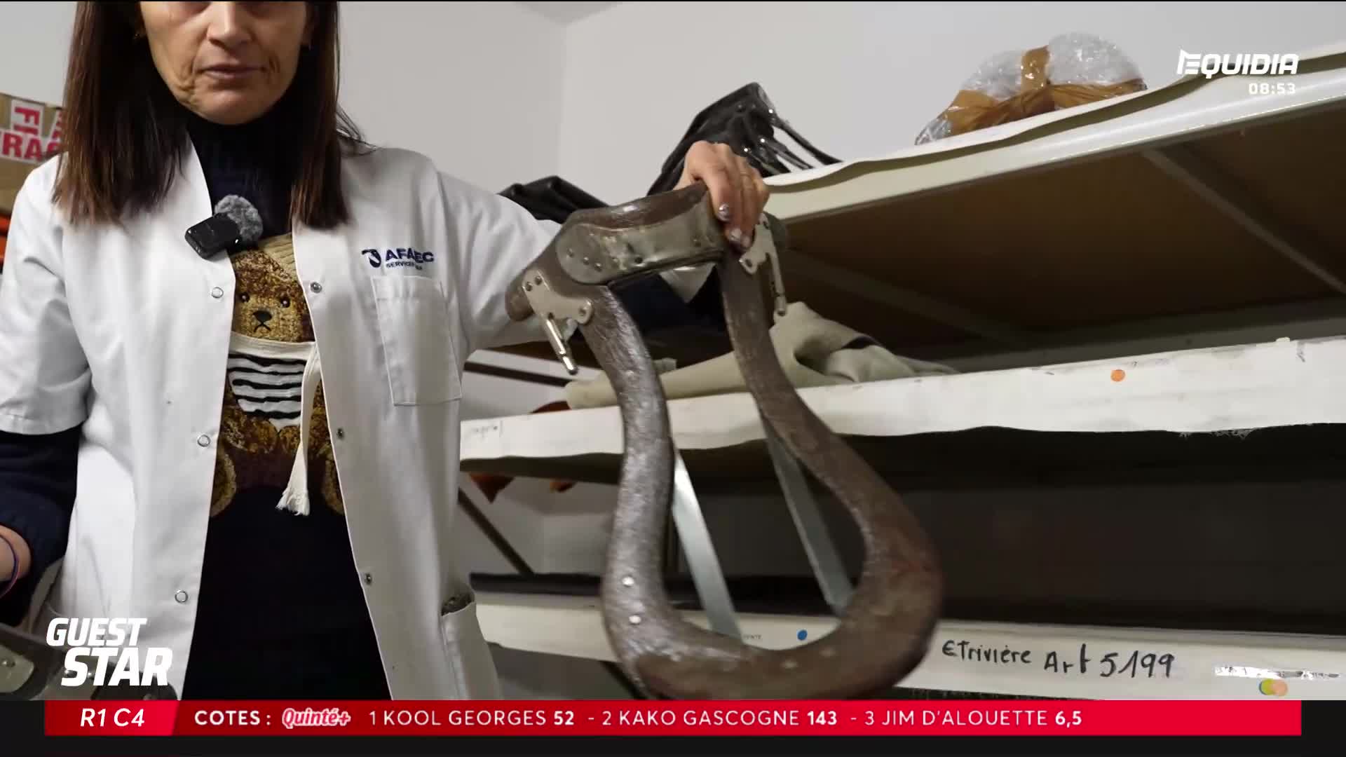 A woman holds up a heavy, curved piece of metal, its surface worn and dark. Behind her, shelves are stacked with what look like saddle parts. A woman holds up a heavy, curved piece of metal, its surface worn and dark. Behind her, shelves are stacked with what look like saddle parts.
