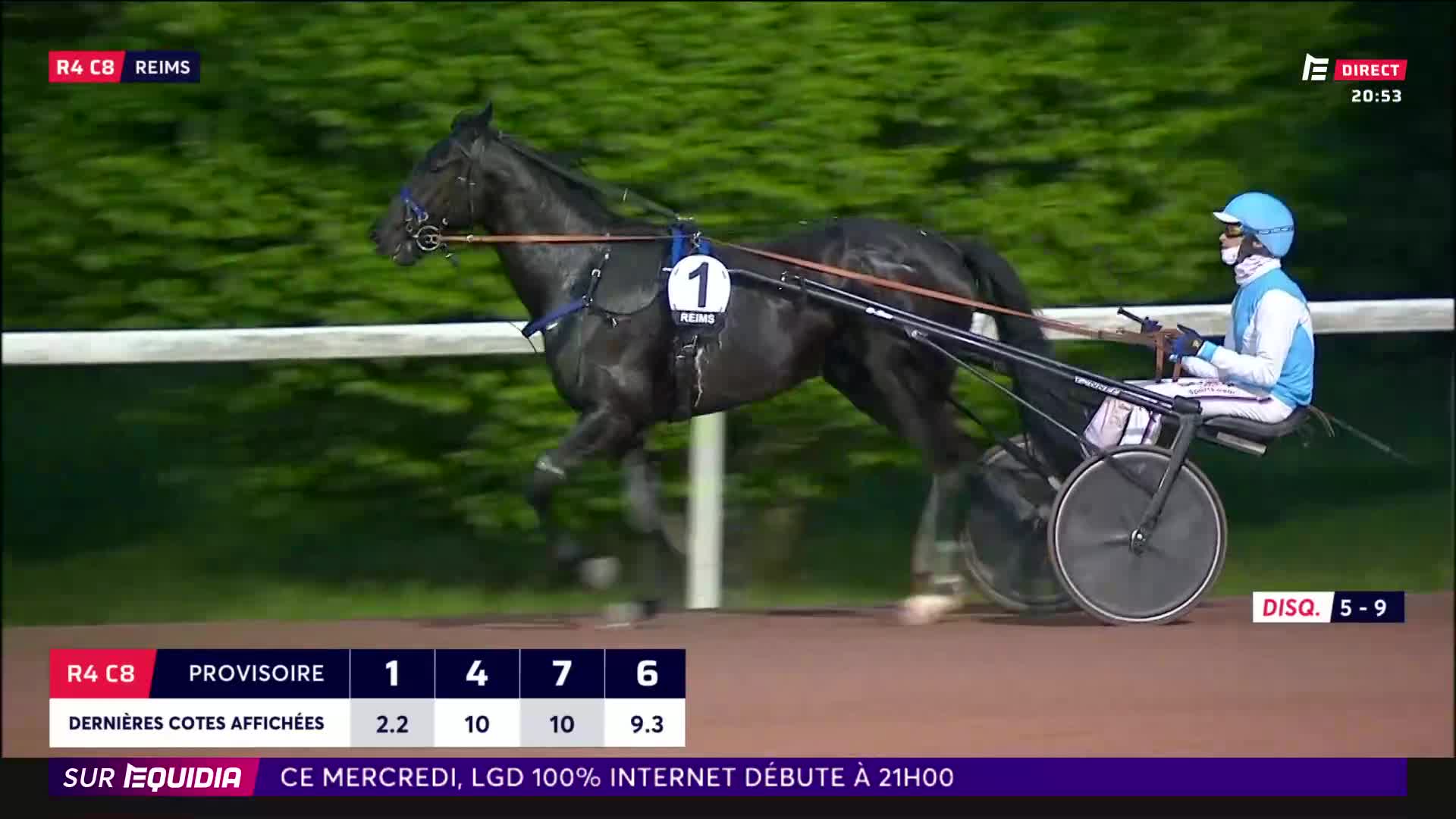 A dark horse pulls a sulky with a driver wearing a blue helmet and silks. The horse's legs are a blur of motion as it races along the track.