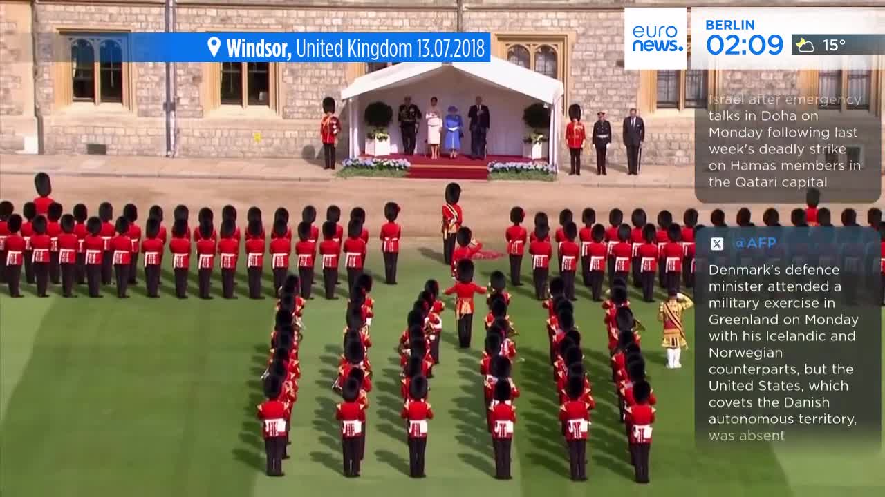 Rows of soldiers in bright red jackets and tall black hats stand at attention on the green. Up on a raised platform, a small group of people, including the Queen, are watching the ceremony.
