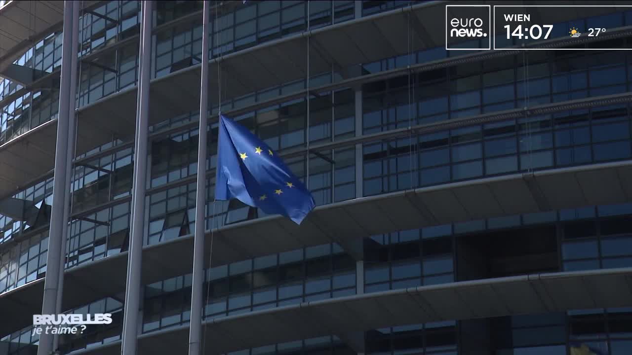 A blue flag with yellow stars hangs limp from a pole, its fabric rippling slightly. The flag is set against a modern building, with the Euronews logo visible in the corner.

