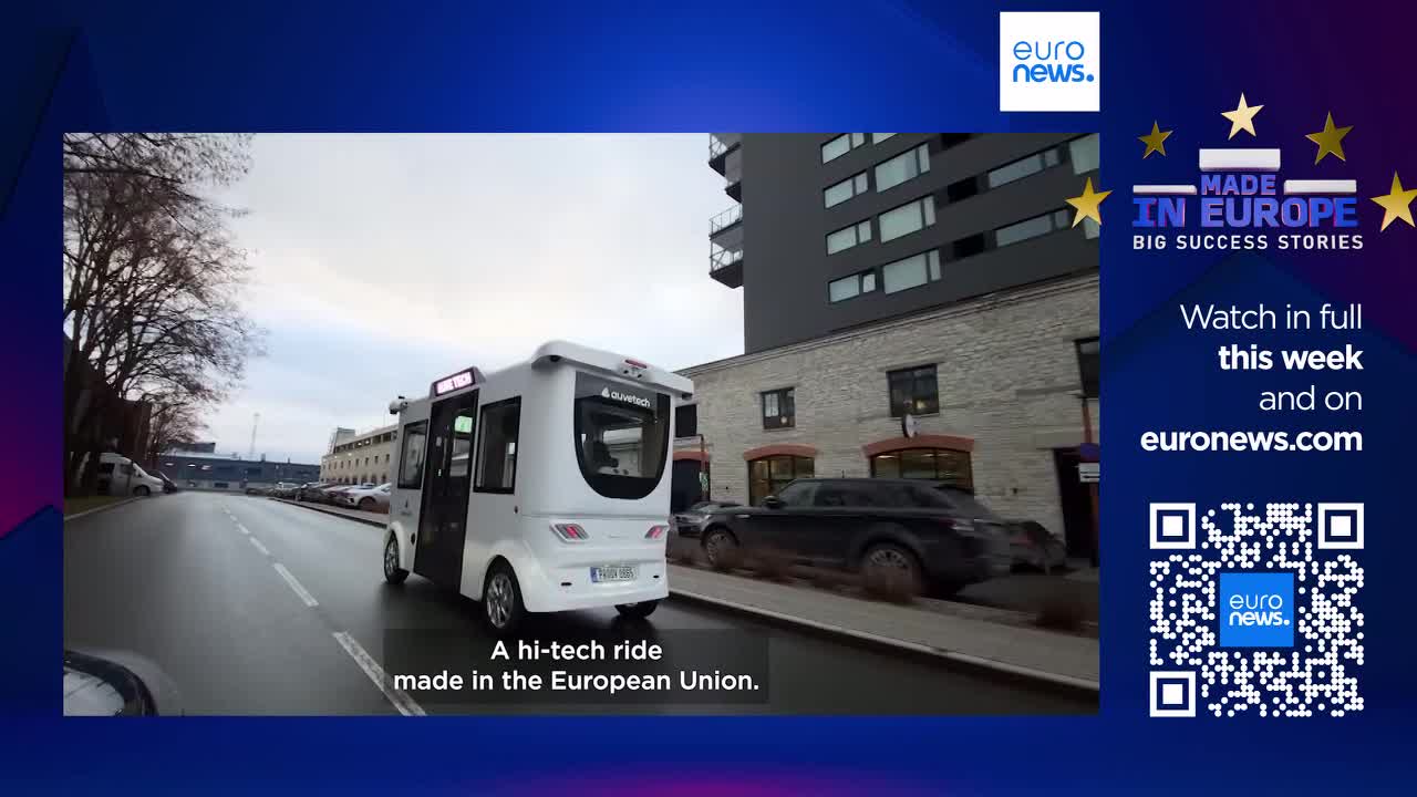 A sleek, white autonomous shuttle glides down a city street. A dark car passes it, its wheels kicking up a bit of spray on the wet pavement. A sleek, white autonomous shuttle glides down a city street. A dark car passes it, its wheels kicking up a bit of spray on the wet pavement.