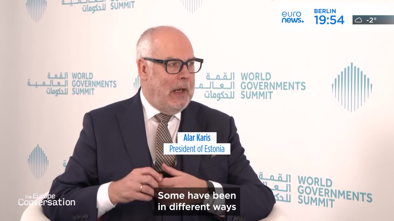 The President of Estonia, Alar Karis, is speaking at the World Governments Summit. He's wearing a dark suit and tie, his hands clasped in front of him. The backdrop features the summit's logo in Arabic and English. The President of Estonia, Alar Karis, is speaking at the World Governments Summit. He's wearing a dark suit and tie, his hands clasped in front of him. The backdrop features the summit's logo in Arabic and English.