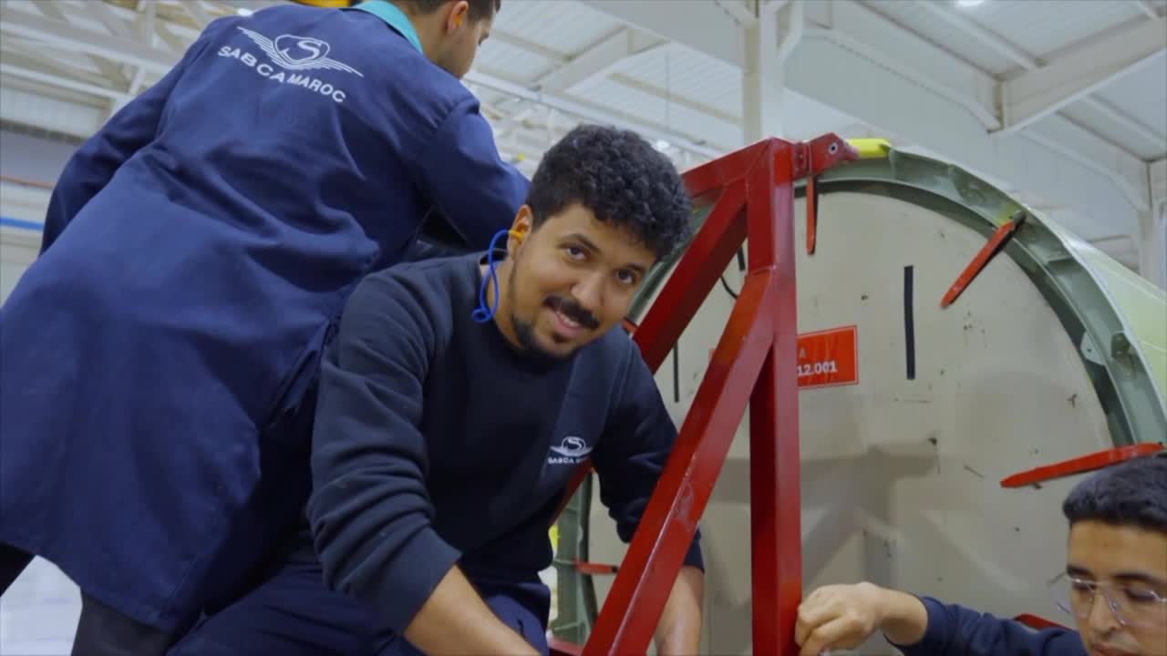 Workers in blue jumpsuits are assembling a large aircraft component. A young man with earplugs smiles at the camera while leaning against a red support structure. Workers in blue jumpsuits are assembling a large aircraft component. A young man with earplugs smiles at the camera while leaning against a red support structure.