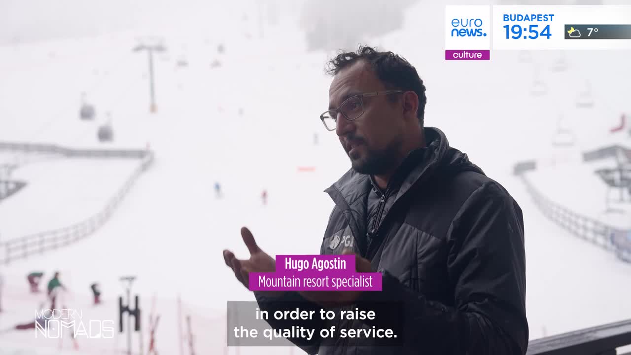 A man speaks with his hands gesturing, while skiers descend a snowy slope behind him. Cable cars glide overhead, carrying people up the mountain. A man speaks with his hands gesturing, while skiers descend a snowy slope behind him. Cable cars glide overhead, carrying people up the mountain.