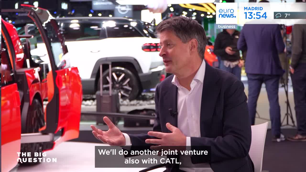 A man in a suit speaks animatedly, gesturing with his hands. Behind him, a bright red car with its doors open sits on display next to a white SUV. A man in a suit speaks animatedly, gesturing with his hands. Behind him, a bright red car with its doors open sits on display next to a white SUV.